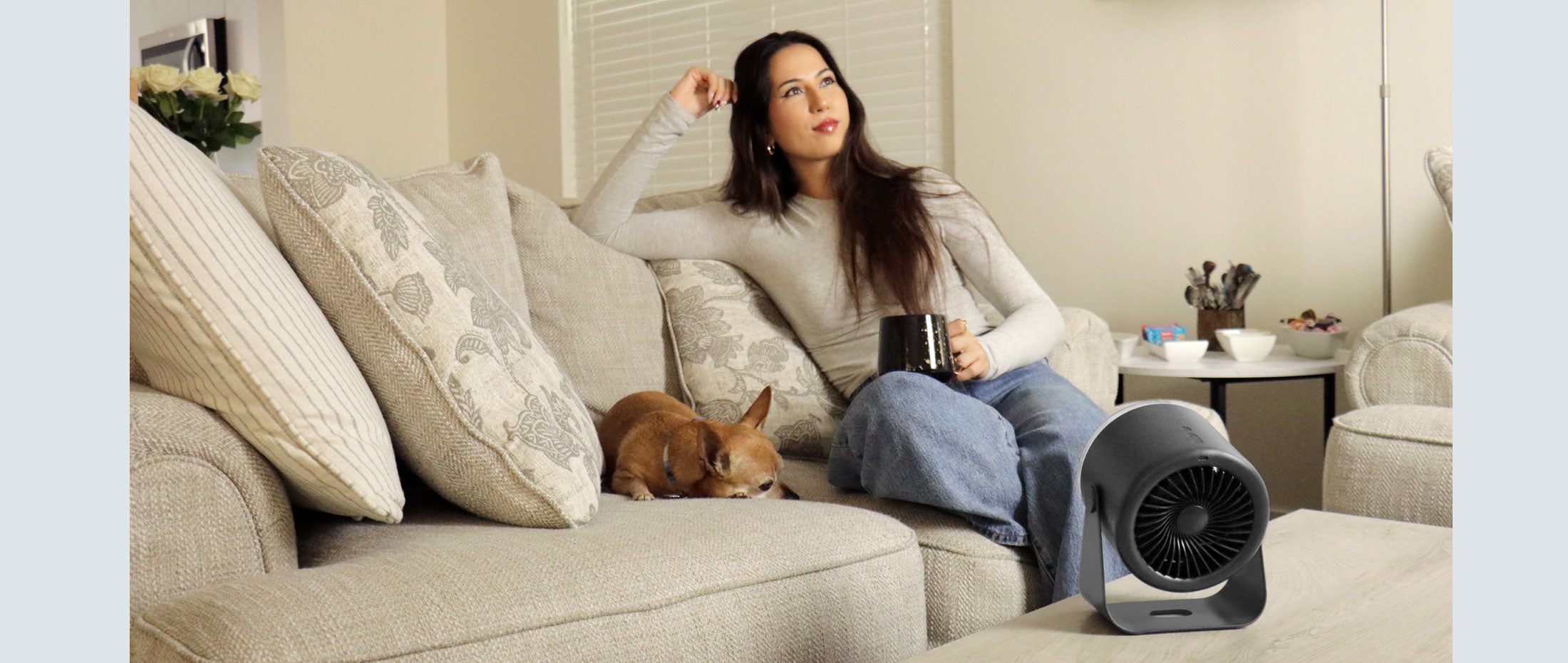 Woman sitting on a couch with a dog and a portable Aluratek fan, in a home setting.